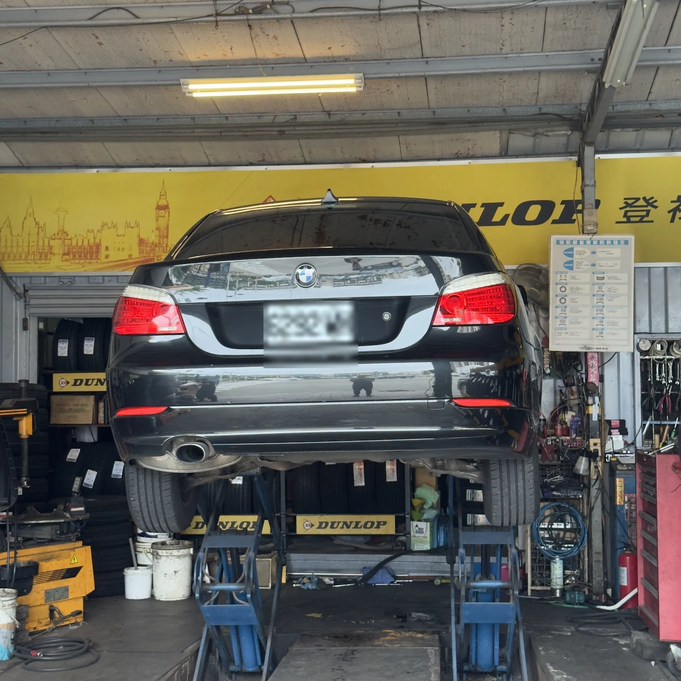 黑色 BMW E60 520d 被架在輪胎行的舉升機上,從車尾方向拍攝