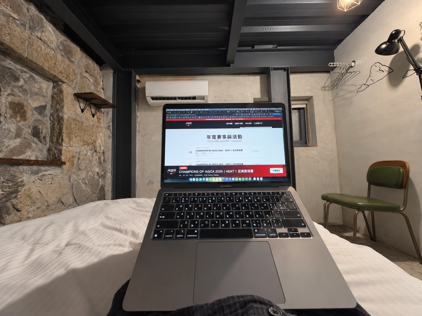 從床上視角拍攝的 MacBook,電腦放在身前,背景是房內石牆、冷氣與掛衣處